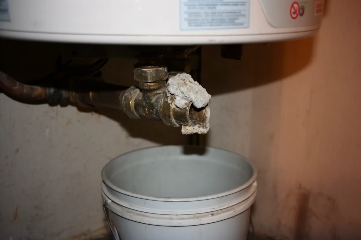 Amateur photo of a water heater drain valve with a bucket under it and mineral scale on the fitting